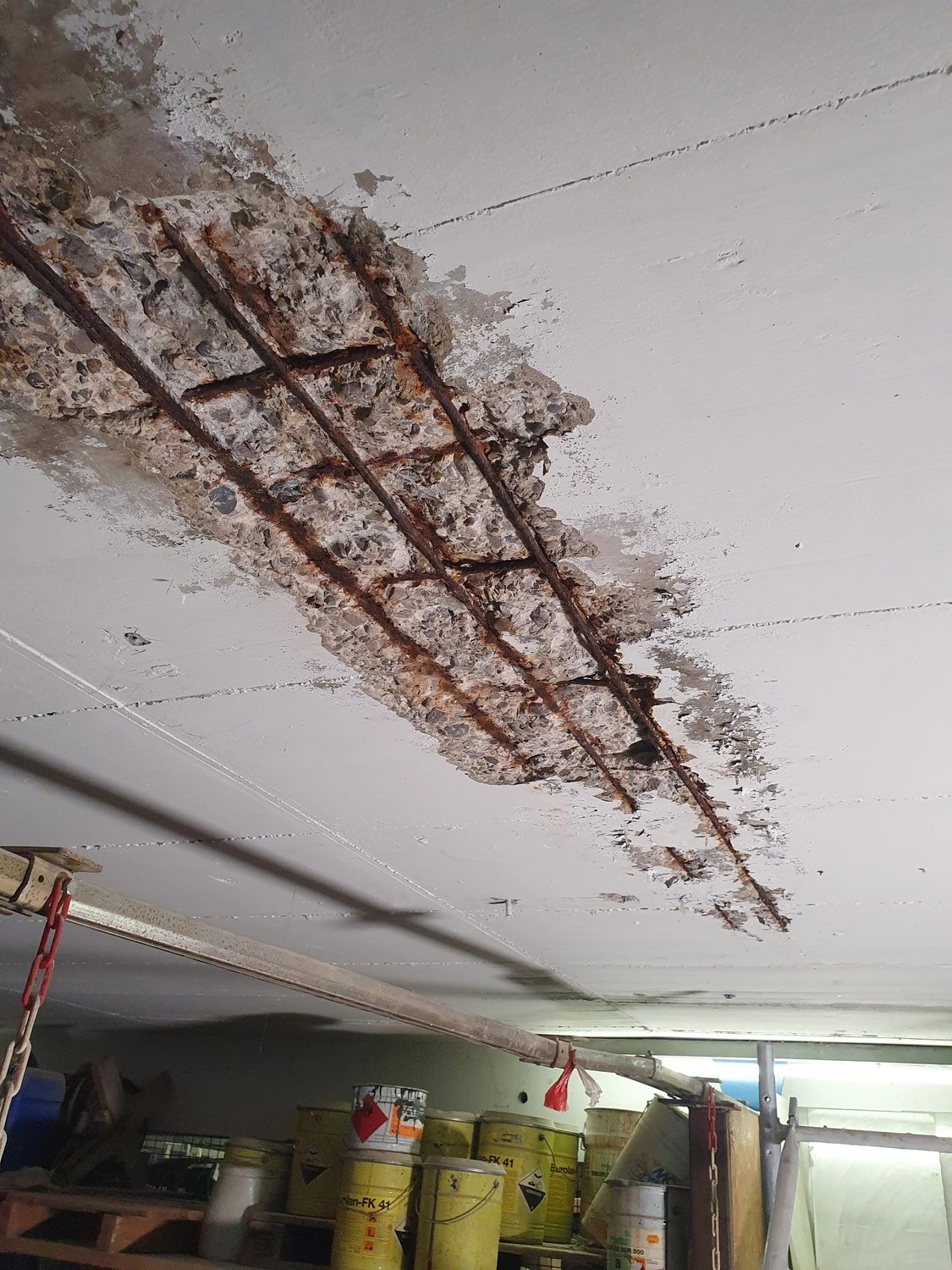 A&S Armbruster & Sieber GmbH - Experten im Bereich Bautenschutz und Bodenbeschichtungen - Decke mit freiliegenden verrosteten Stahlbewehrungsstäben und beschädigtem Beton in einem Lagerraum mit gelben Fässern und Baumaterialien