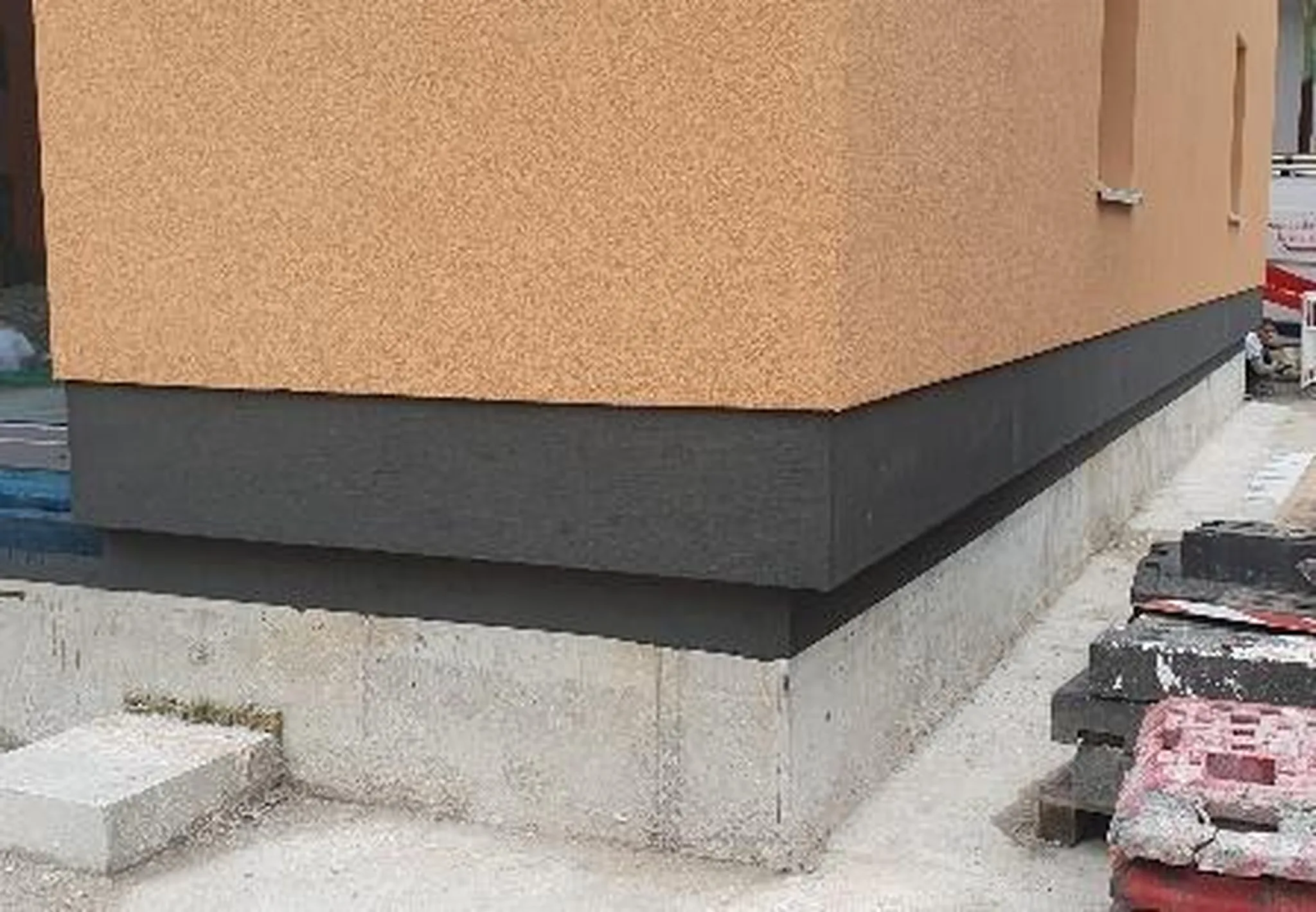 A&S Armbruster & Sieber GmbH - Experten im Bereich Bautenschutz und Bodenbeschichtungen - Ecke eines im Bau befindlichen Gebäudes mit beiger Oberwand, schwarzem Mittelteil und Betonfundament, in der Nähe Baumaterialien.