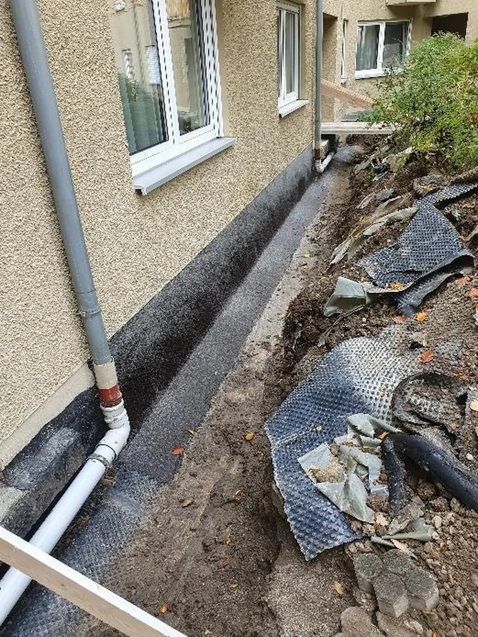 A&S Armbruster & Sieber GmbH - Experten im Bereich Bautenschutz und Bodenbeschichtungen - Baustelle neben einem beigen Stuckgebäude mit einem entlang des Fundaments ausgehobenen Graben, schwarzem Abdichtungsmaterial auf der Grundmauer und installierten Entwässerungsrohren.