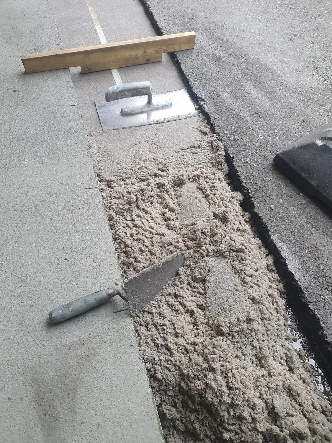 A&S Armbruster & Sieber GmbH - Experten im Bereich Bautenschutz und Bodenbeschichtungen - Frisch aufgetragener Mörtel zwischen Betongehweg und Asphaltstraße mit zwei Metallkellen und einem darauf liegenden Holzestrichwerkzeug