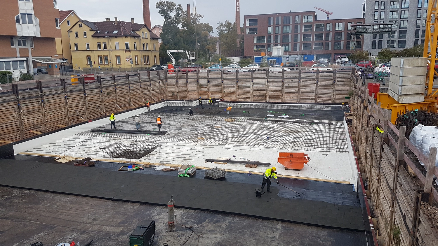 A&S Armbruster & Sieber GmbH - Experten im Bereich Bautenschutz und Bodenbeschichtungen - Baustelle mit Arbeitern, die Bewehrungsstäbe und Abdichtungsbahnen in einer tiefen Baugrube installieren, die von hölzernen Stützmauern umgeben ist.