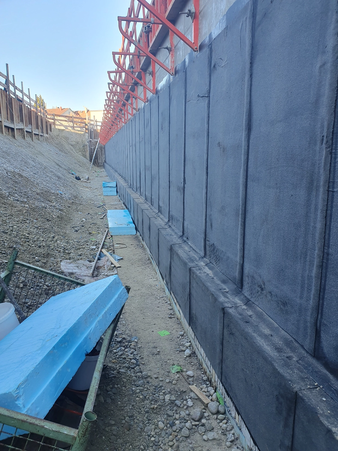 A&S Armbruster & Sieber GmbH - Experten im Bereich Bautenschutz und Bodenbeschichtungen - Baugrube mit hoher Betonstützmauer mit schwarzem Abdichtungsmaterial und darüber liegenden roten Stützwinkeln
