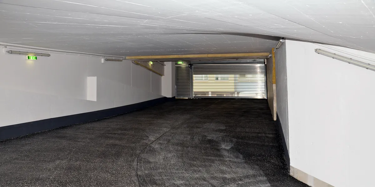 A&S Armbruster & Sieber GmbH - Experten im Bereich Bautenschutz und Bodenbeschichtungen - Geschwungene Parkhaus-Ausfahrtsrampe mit teilweise geschlossenem Metallrolltor und weißen Wänden