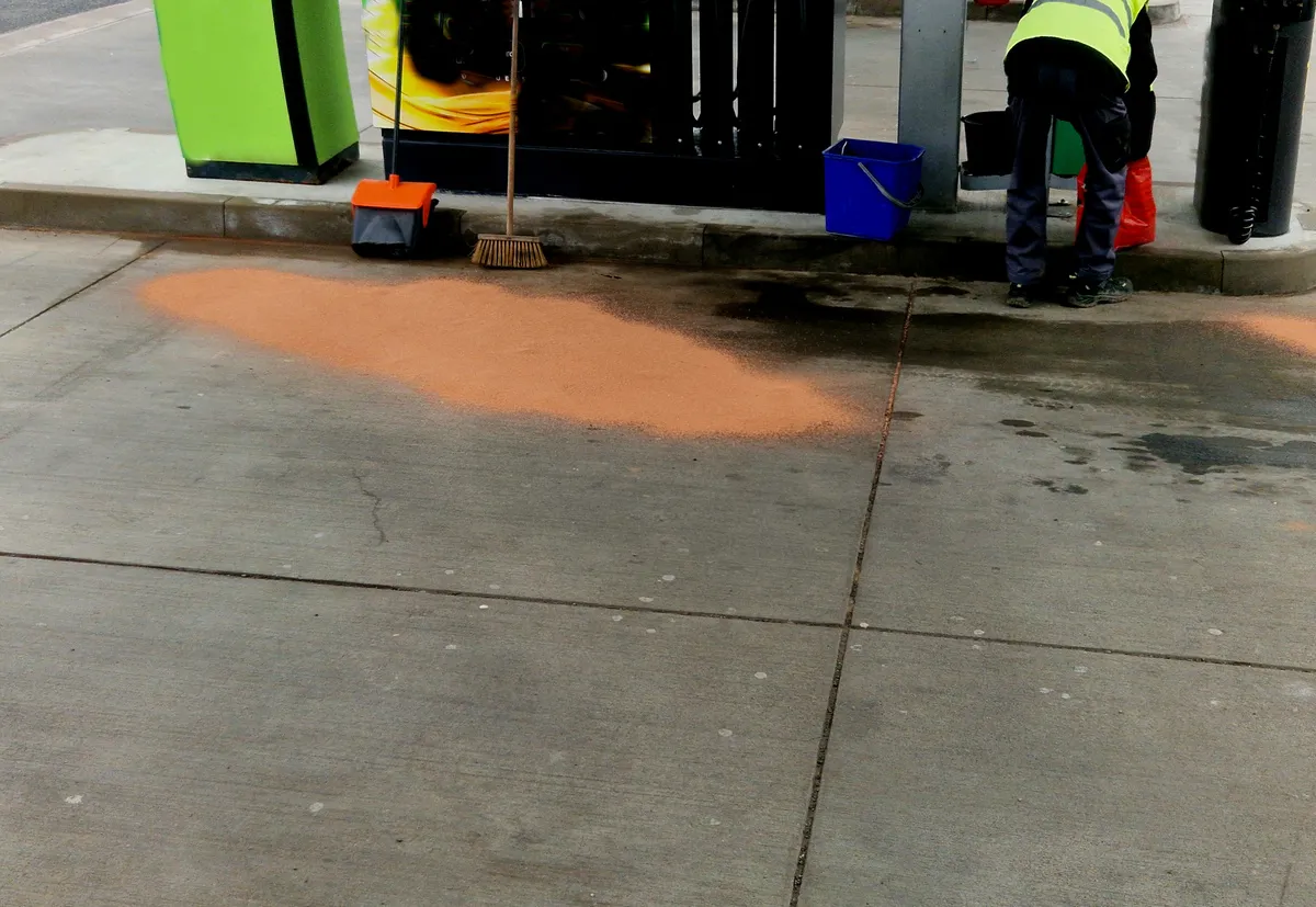 A&S Armbruster & Sieber GmbH - Experten im Bereich Bautenschutz und Bodenbeschichtungen - Person in Sicherheitsweste reinigt einen Ölteppich an einer Tankstelle mit absorbierendem orangefarbenem Pulver und Reinigungswerkzeugen in der Nähe