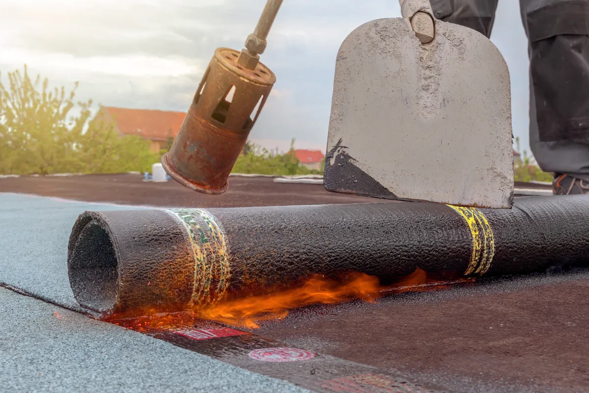 A&S Armbruster & Sieber GmbH - Experten im Bereich Bautenschutz und Bodenbeschichtungen - Arbeiter trägt mit Fackel und Schaufel ein gerolltes Dachmaterial auf und erhitzt es, wobei darunter Flammen sichtbar sind